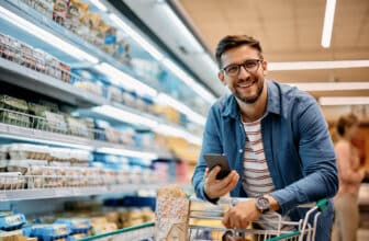 Happy man using mobile phone app while buying groceries in supermarket and looking at camera.