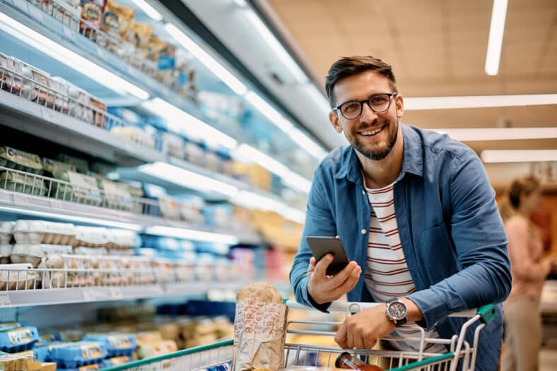 Happy man using mobile phone app while buying groceries in supermarket and looking at camera.